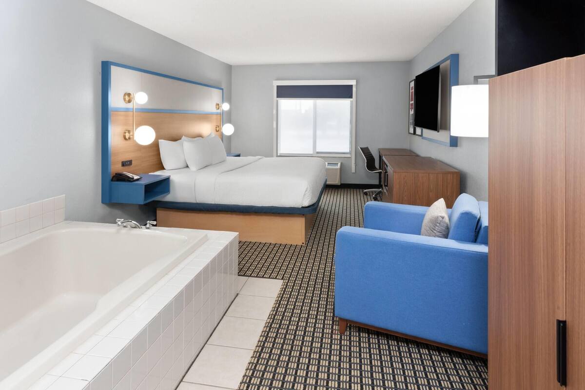 A modern hotel room with a large bed, a separate sofa, a bathtub, and a flat-screen TV mounted on the wall.