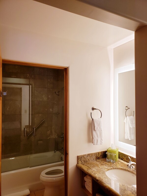 Un baño con ducha de vidrio, inodoro blanco, lavamanos con encimera de mármol y espejo sobre el lavamanos.