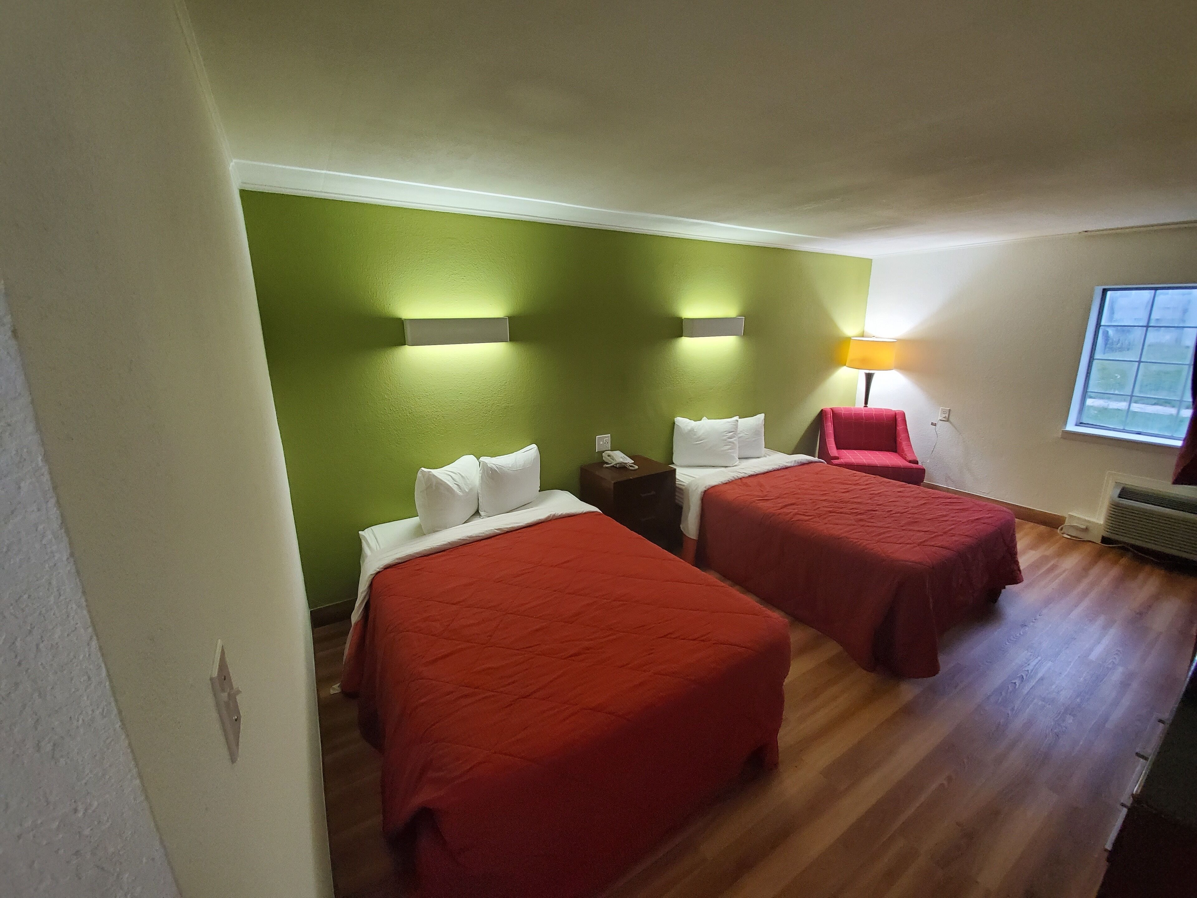 Een hotelkamer met twee bedden, groene muren, een rode stoel, een nachtkastje en een raam.
