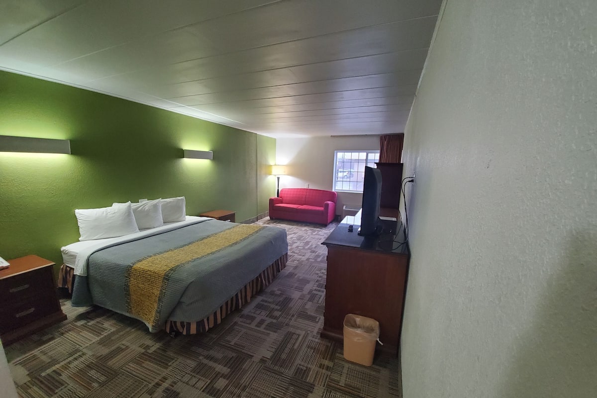 A hotel room with a bed, a desk with a computer, a red chair, and a window with a view of the street.