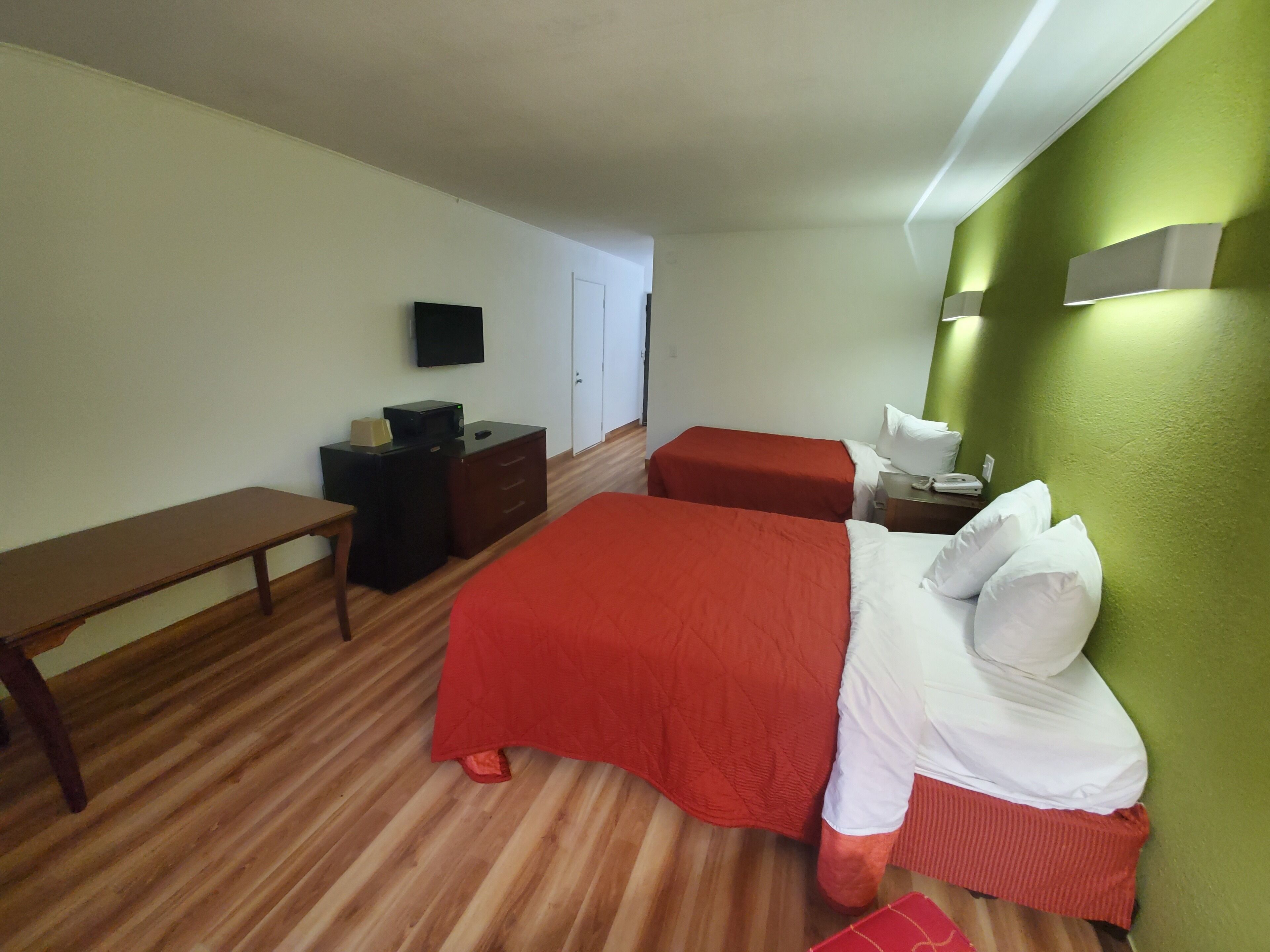 A hotel room with two beds, a desk, a bench, a flat-screen TV, and a green accent wall.