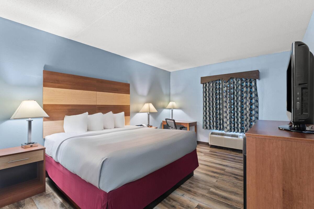 A hotel room with a large bed, two bedside tables with lamps, a desk with a chair, a television, and a window with patterned curtains.