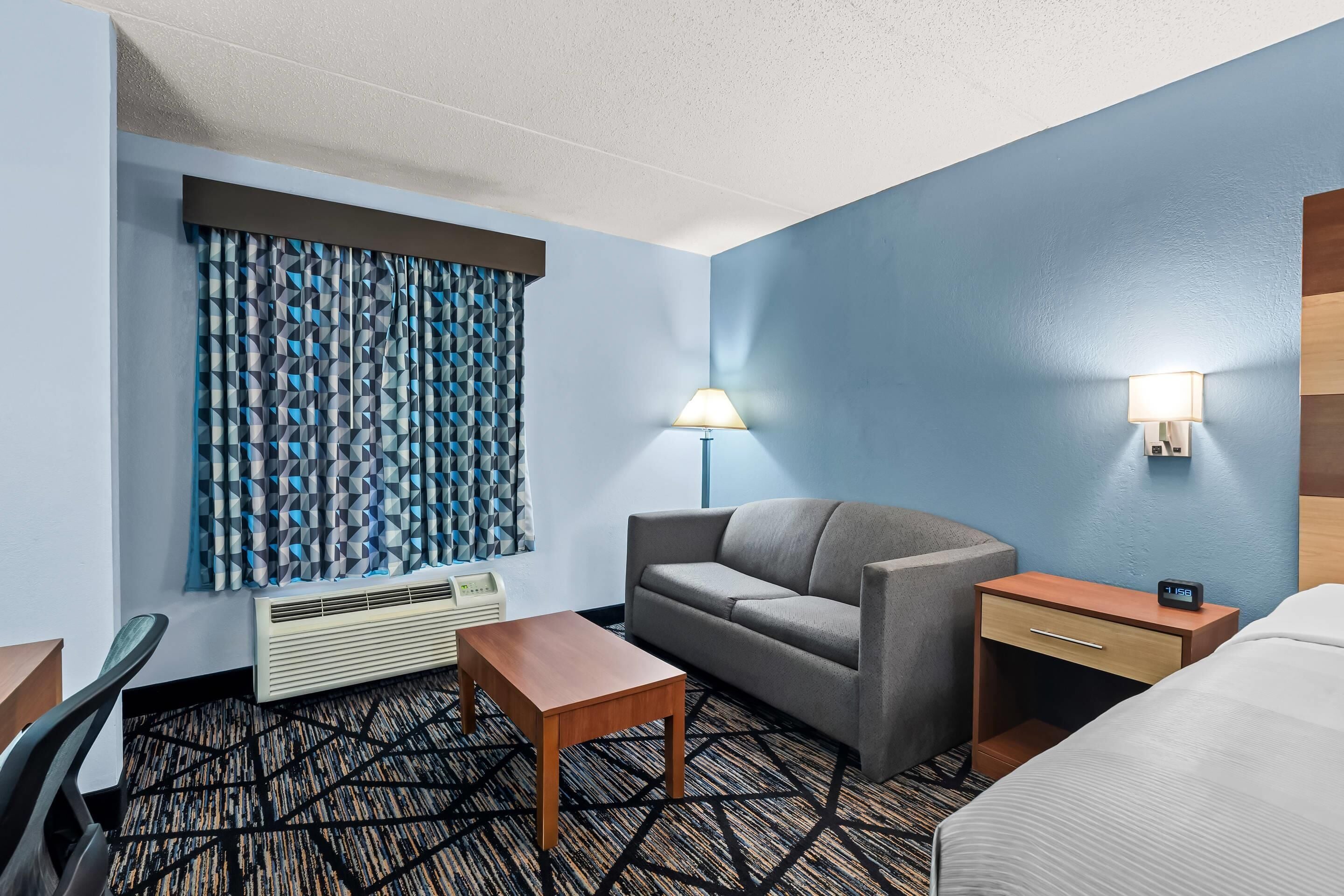 Une chambre d’hôtel avec un canapé, une petite table, une lampe, une table de chevet avec une horloge et un lit.