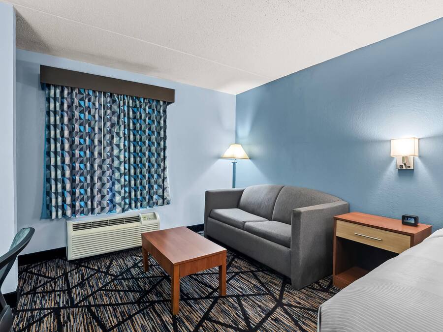 Habitación de hotel con sofá, una mesita, lámpara, mesita de noche con reloj y cama.