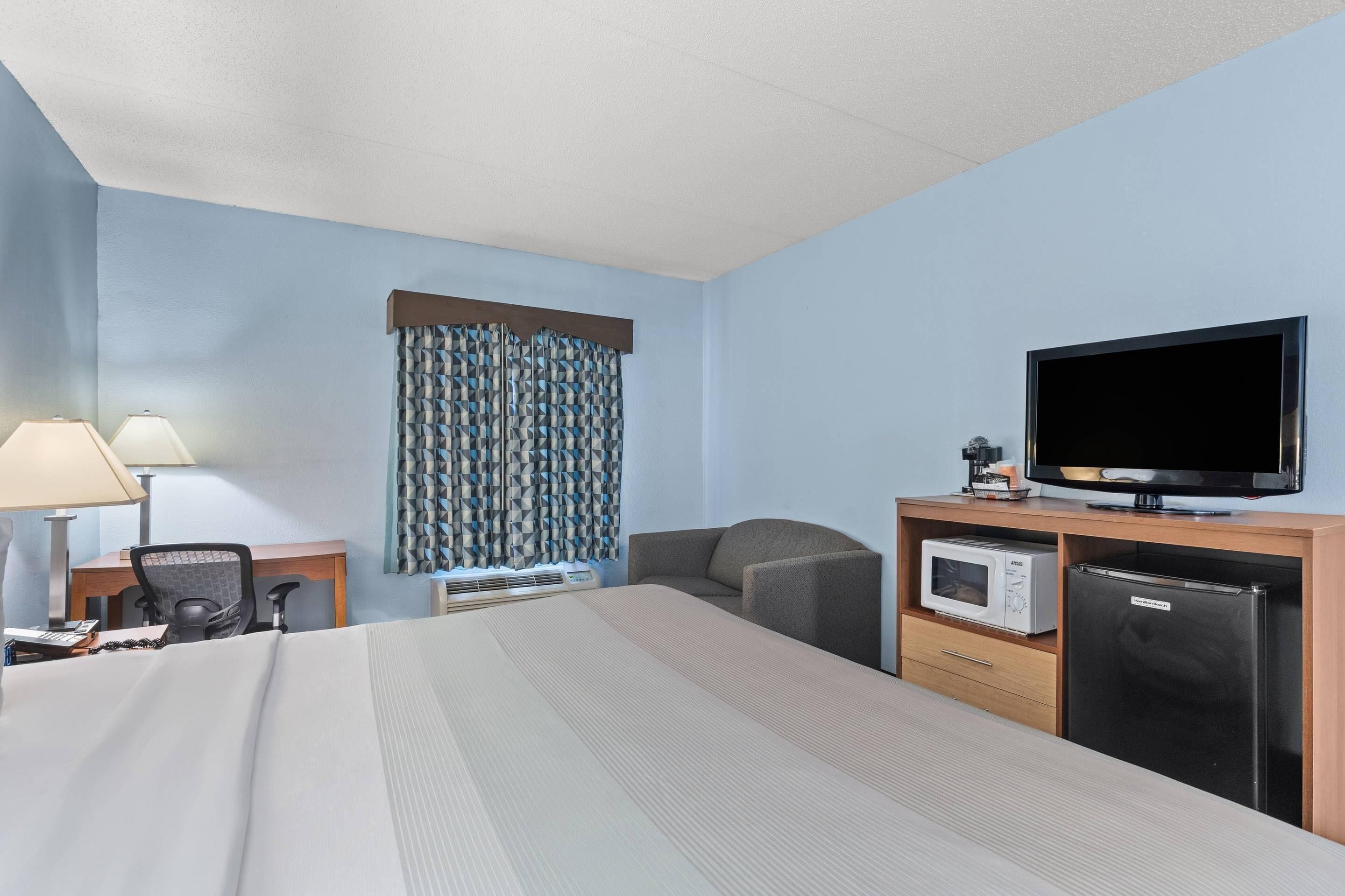 Ett hotellrum med en säng, ett skrivbord med en lampa, en stol, en tv, en mikrovågsugn och ett kylskåp.