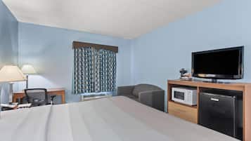 Ett hotellrum med en säng, ett skrivbord med en lampa, en stol, en tv, en mikrovågsugn och ett kylskåp.
