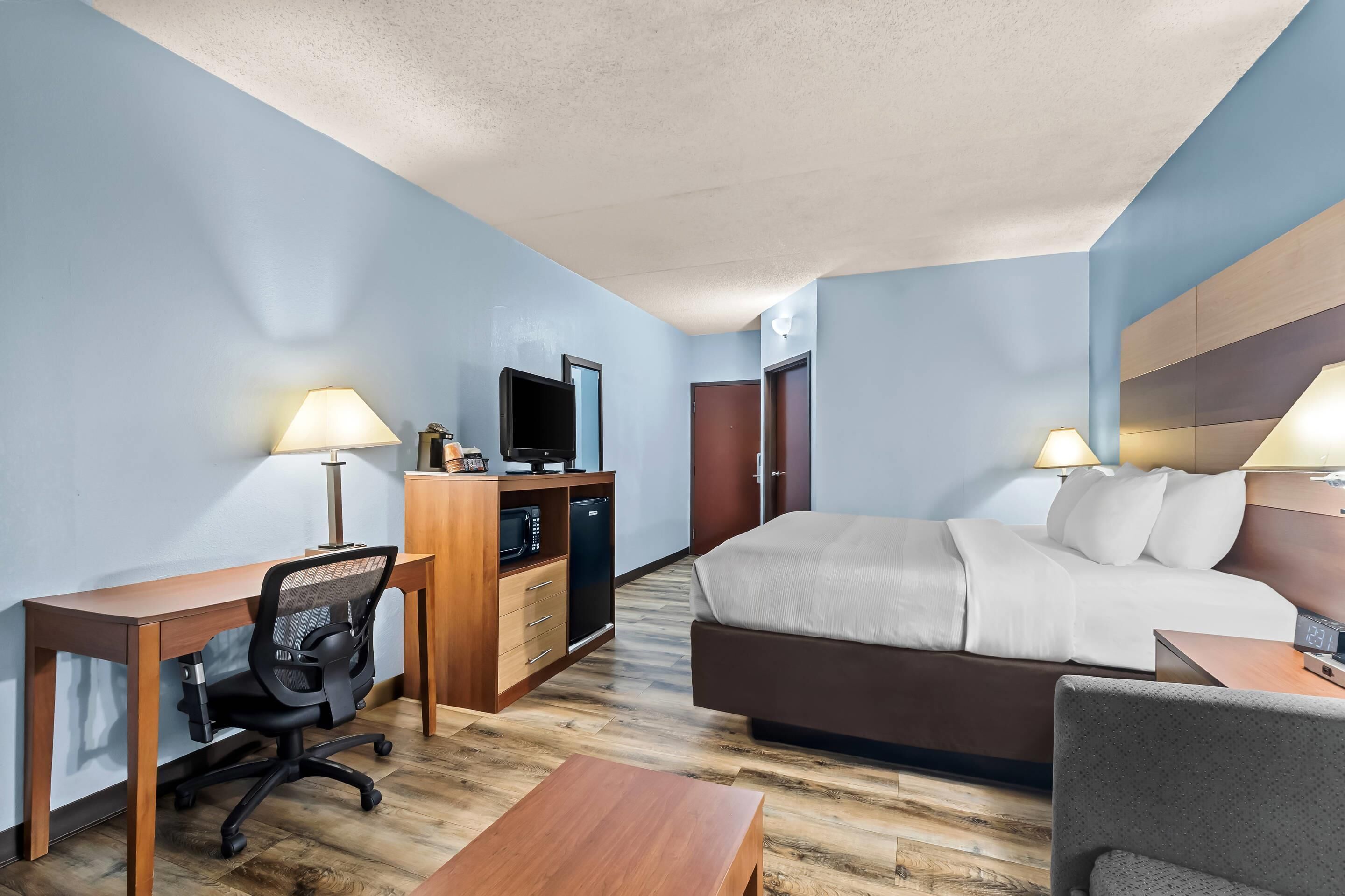 A hotel room with a bed, desk, chair, TV, and a small kitchenette.