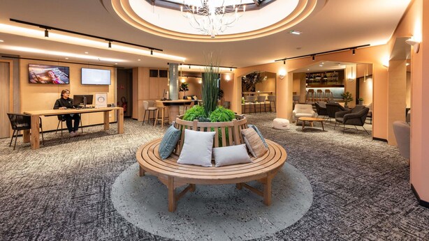 A hotel lobby with a central wooden bench, a skylight, a bar area, and seating arrangements. - Novotel Mâcon Nord – Porte de Bourgogne (Macon)