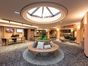 A hotel lobby with a central wooden bench, a skylight, a bar area, and seating arrangements.