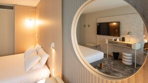 A modern hotel room with a bed, a round mirror, a desk with a chair, and a TV. - Novotel Mâcon Nord – Porte de Bourgogne (Macon)