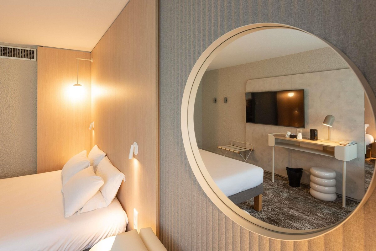 Una habitación de hotel moderna con una cama, un espejo redondo, un escritorio con una silla y un televisor.
