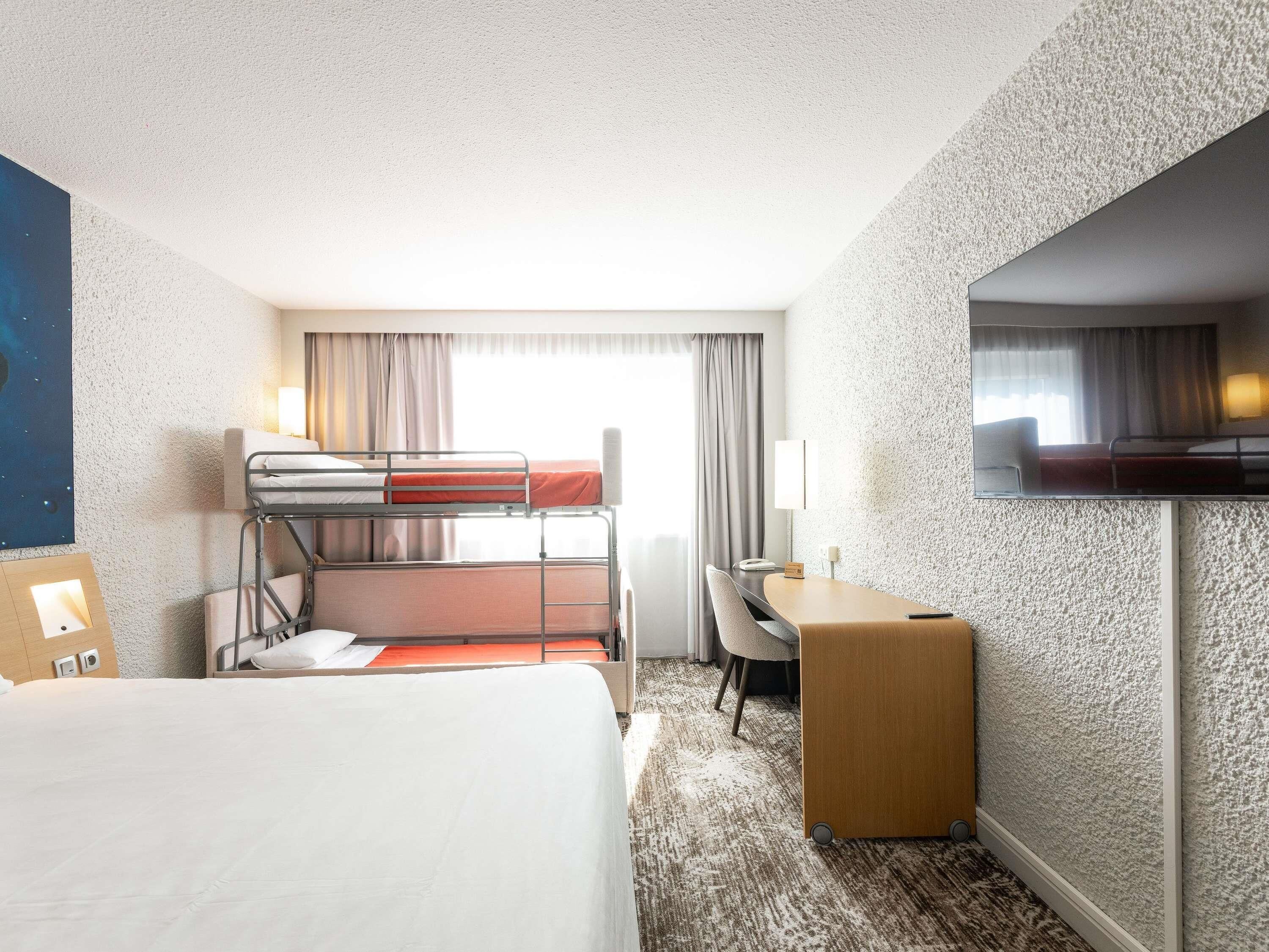 Une chambre d’hôtel avec des lits superposés, un bureau, une chaise, un téléviseur à écran plat et une grande fenêtre avec des rideaux.