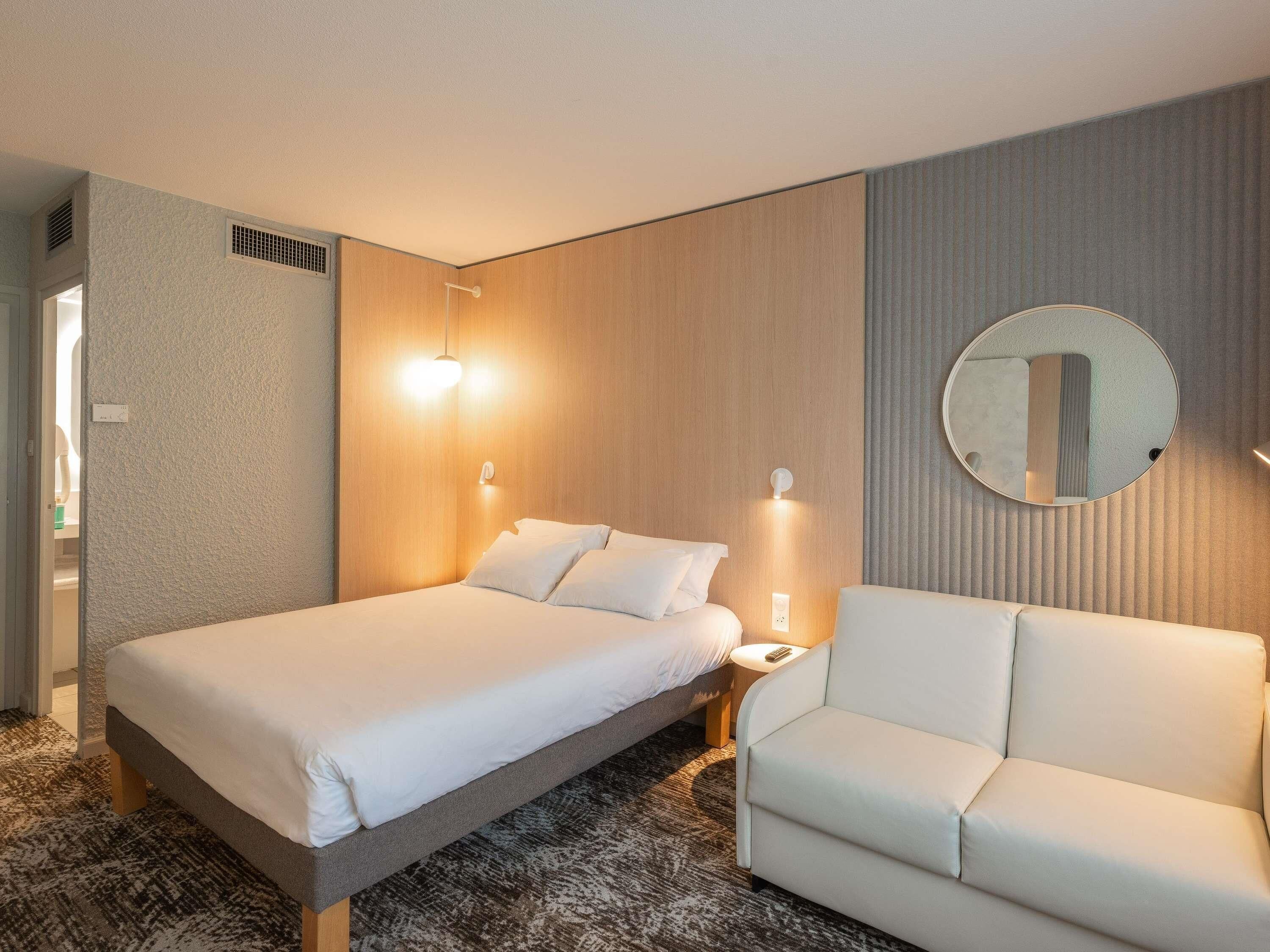 Una habitación de hotel con cama, sofá, espejo redondo e iluminación en la pared.