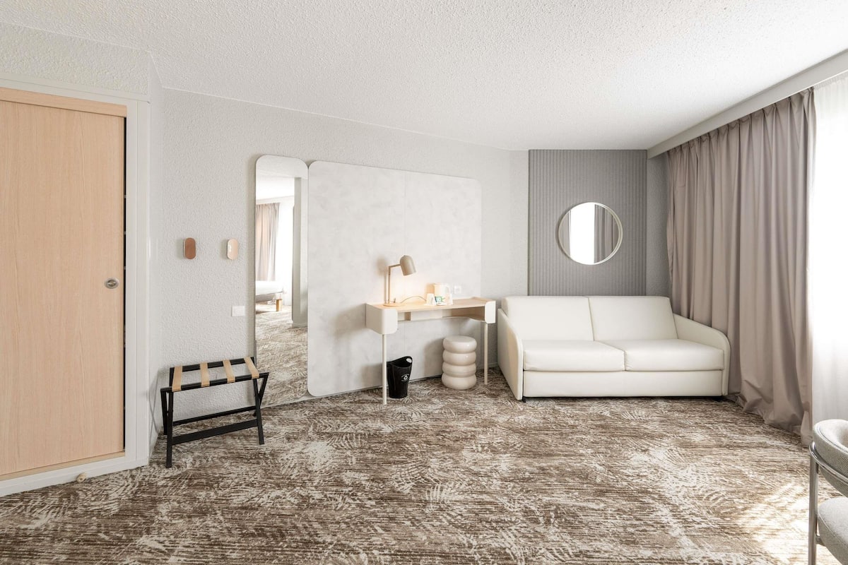 Ein Hotelzimmer mit einer weißen Couch, einem kleinen Tisch mit einer Lampe, einem Spiegel, einem Mülleimer und einem gemusterten Teppich.