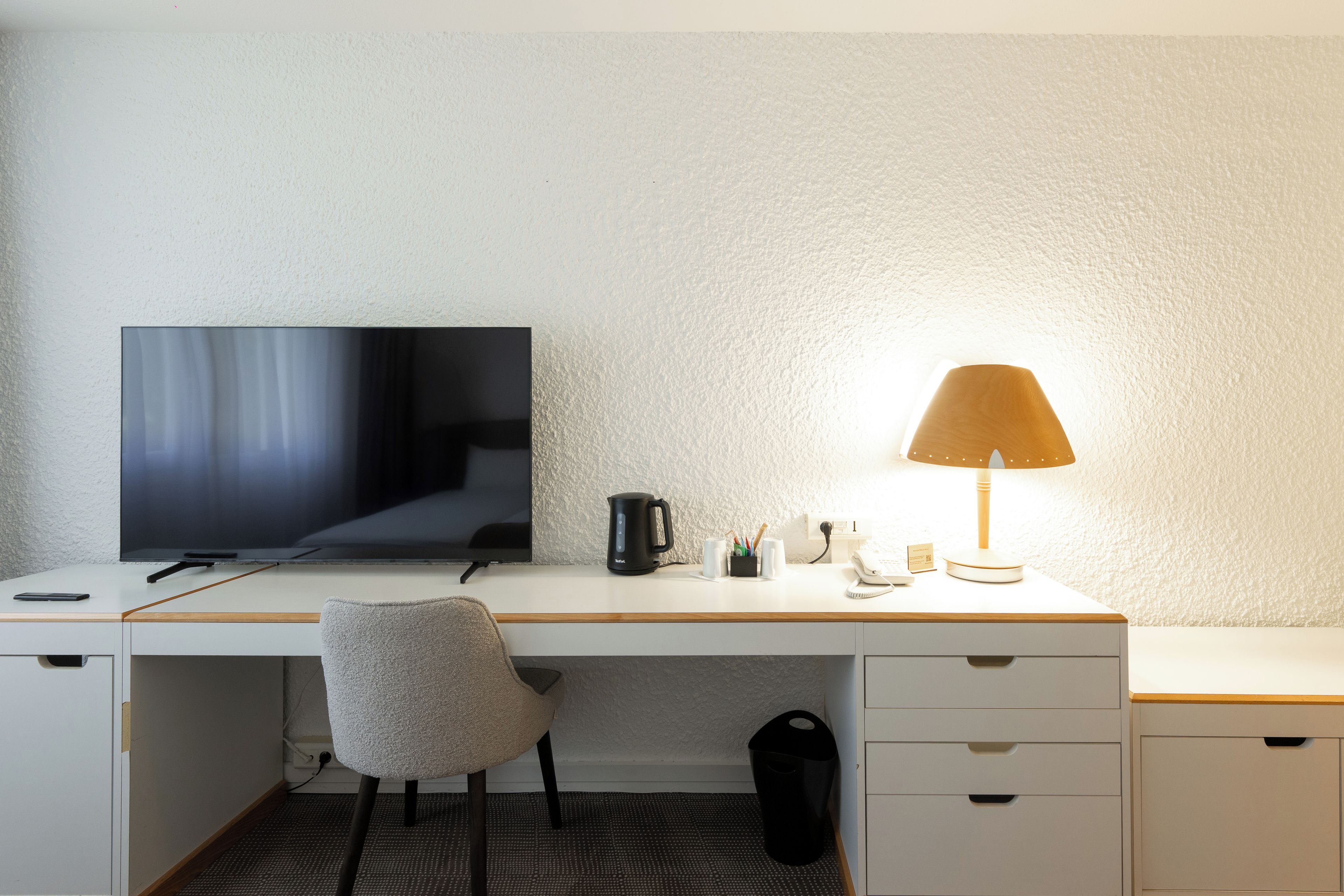 Une chambre d’hôtel équipée d’un téléviseur à écran plat, d’un bureau avec une lampe, d’une chaise, d’une poubelle et d’une petite table sur laquelle se trouvent un téléphone et un bloc-notes.