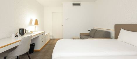 A hotel room with a bed, a desk with a lamp, a chair, a small table, and a sofa.