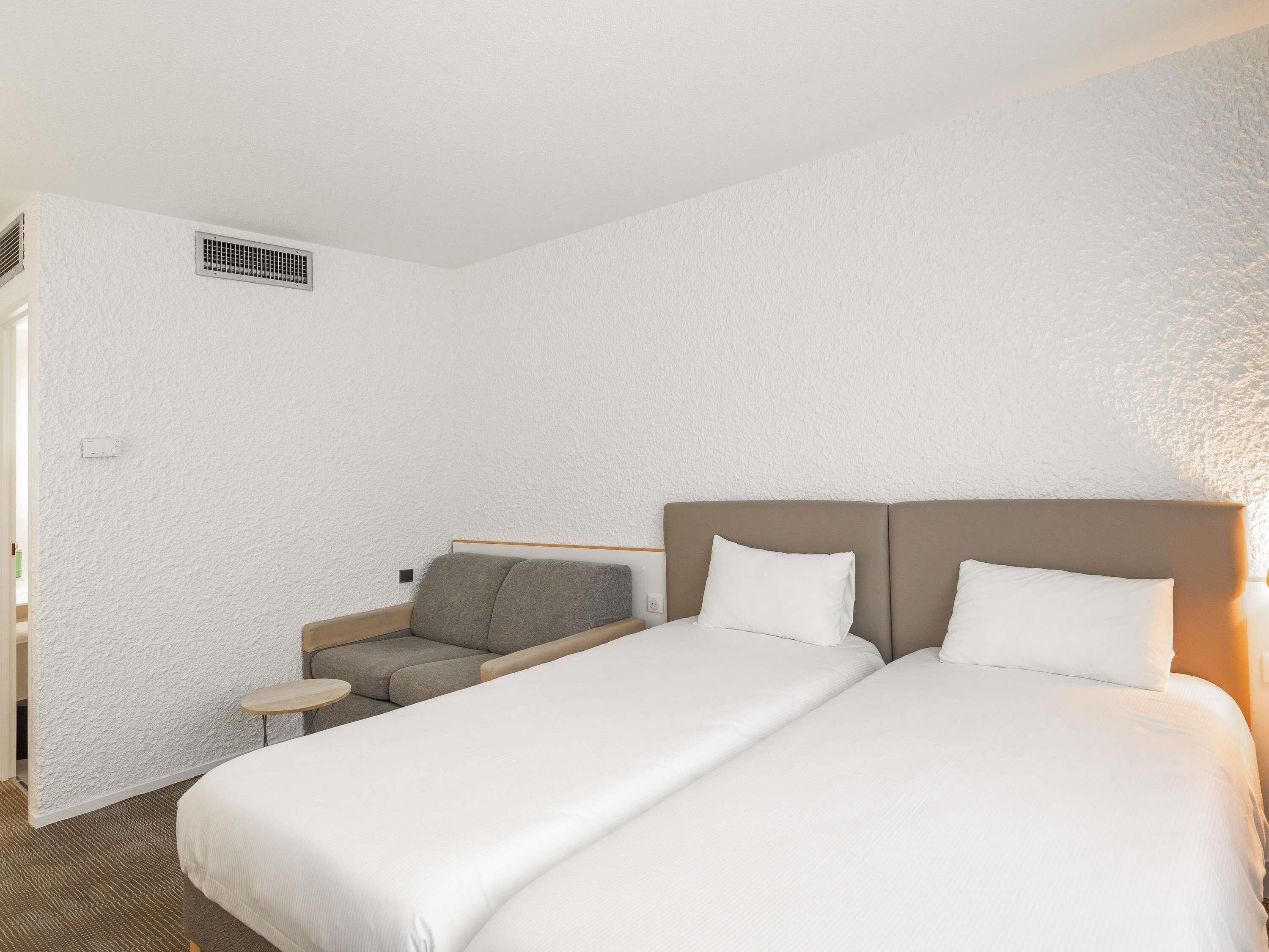 Quarto de hotel com duas camas, um sofá pequeno, uma mesa redonda e um respiradouro fixado na parede.