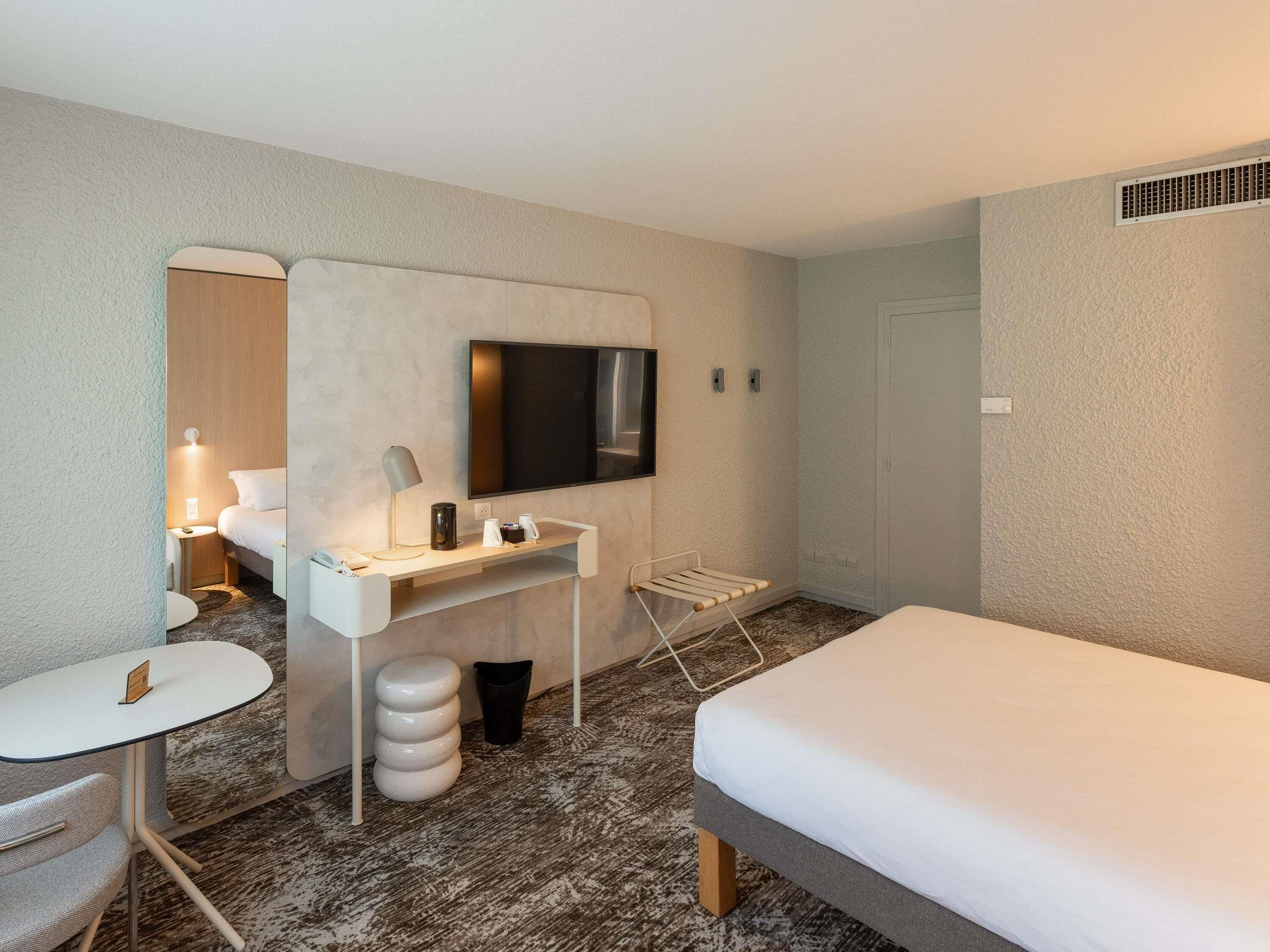 Ett hotellrum med en säng, ett skrivbord med en platt-TV, en stol, ett litet bord och en papperskorg.