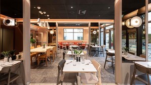 A modern restaurant interior with multiple dining tables and chairs, a central seating area, and large windows providing natural light. - Novotel Mâcon Nord – Porte de Bourgogne (Macon)