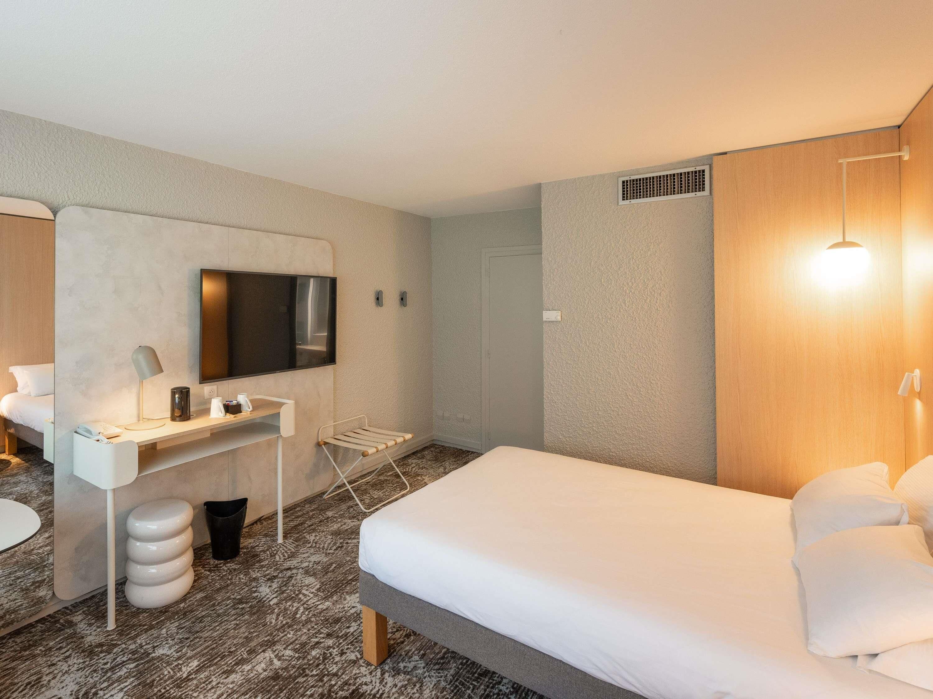 A hotel room with a bed, a desk with a flat-screen TV, a chair, a small table, and a wall-mounted shelf.