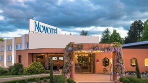 A hotel with a sign reading 'NOVOTEL,' a covered entrance with steps, and a garden area with hedges and shrubs. - Novotel Mâcon Nord – Porte de Bourgogne (Macon)