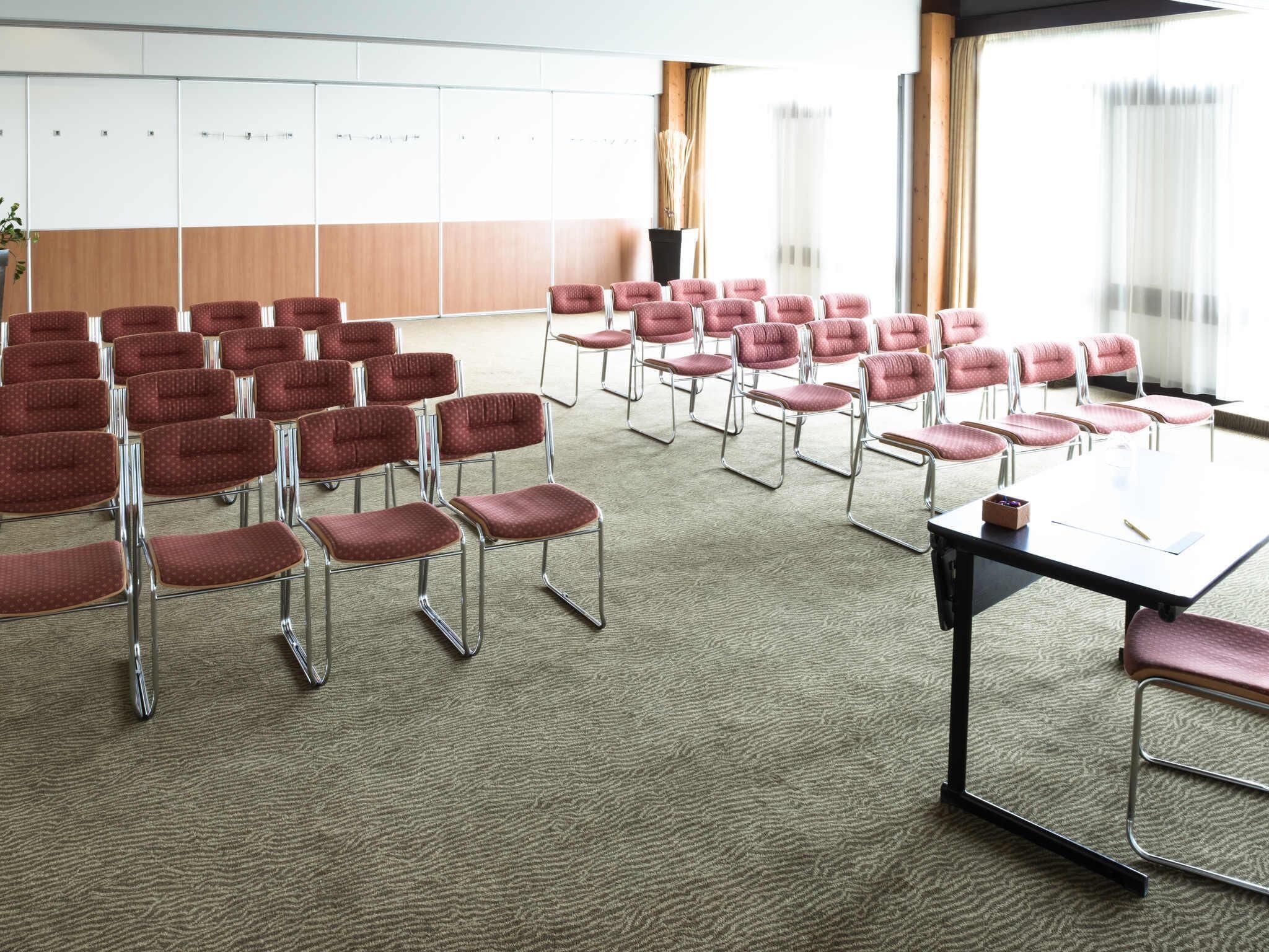 A conference room with rows of red chairs, a large table, and a view of the outdoors through large windows.