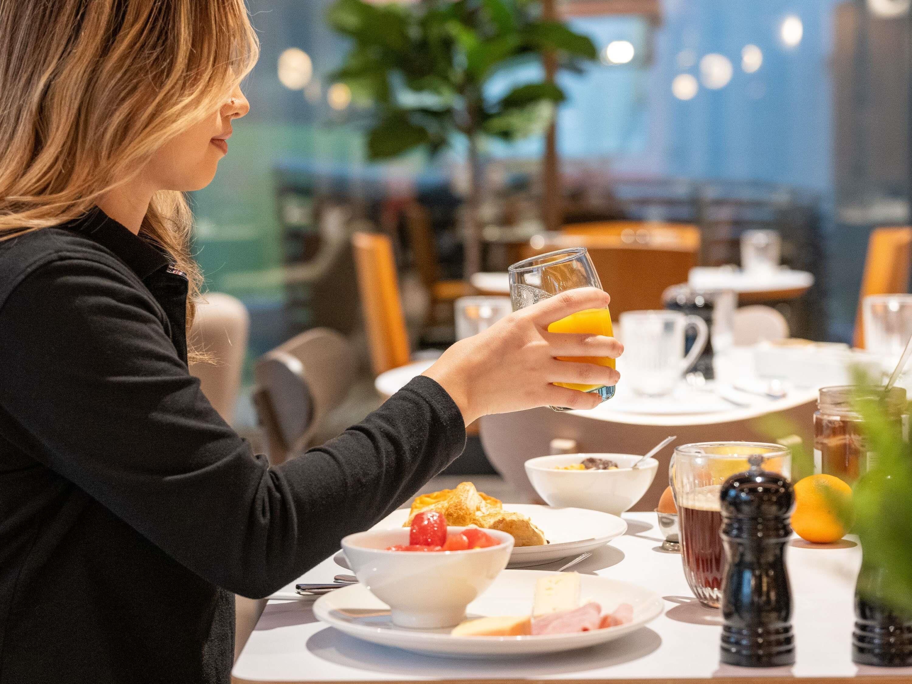 A person holding a glass of orange juice at a breakfast table with various food items and a salt shaker.