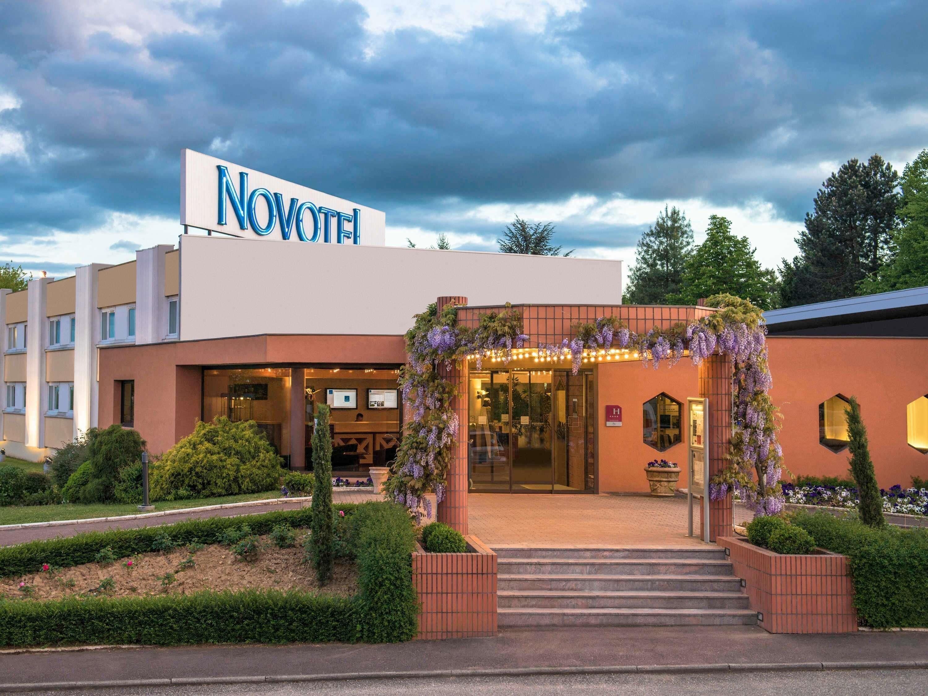 Un hôtel avec une enseigne indiquant « NOVOTEL », une entrée couverte avec des marches et un espace jardin agrémenté de haies et d’arbustes.