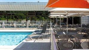 Zona junto a la piscina con sillones de descanso y sombrillas, piscina y un edificio con tejado de baldosas al fondo.