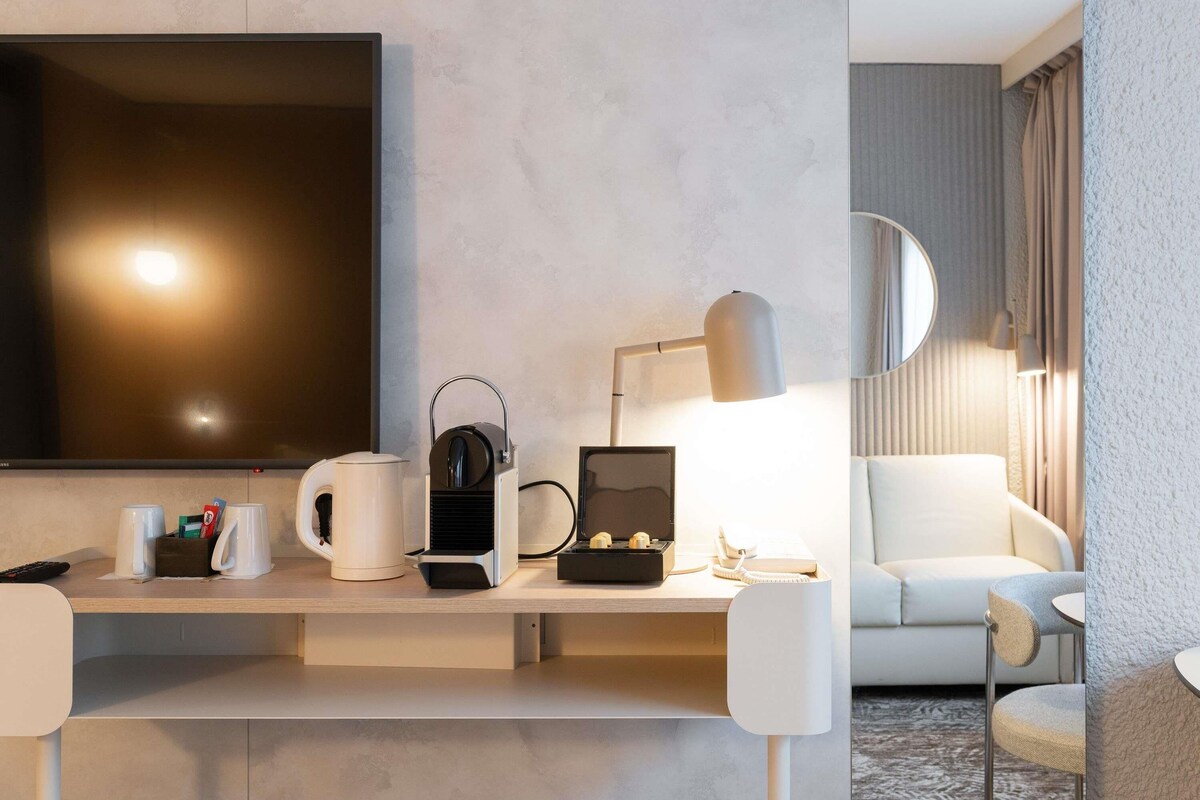Habitación de hotel moderna con un televisor de pantalla plana grande, un escritorio con lámpara, cafetera y una caja de seguridad pequeña. Se observa un sofá y una silla al fondo.