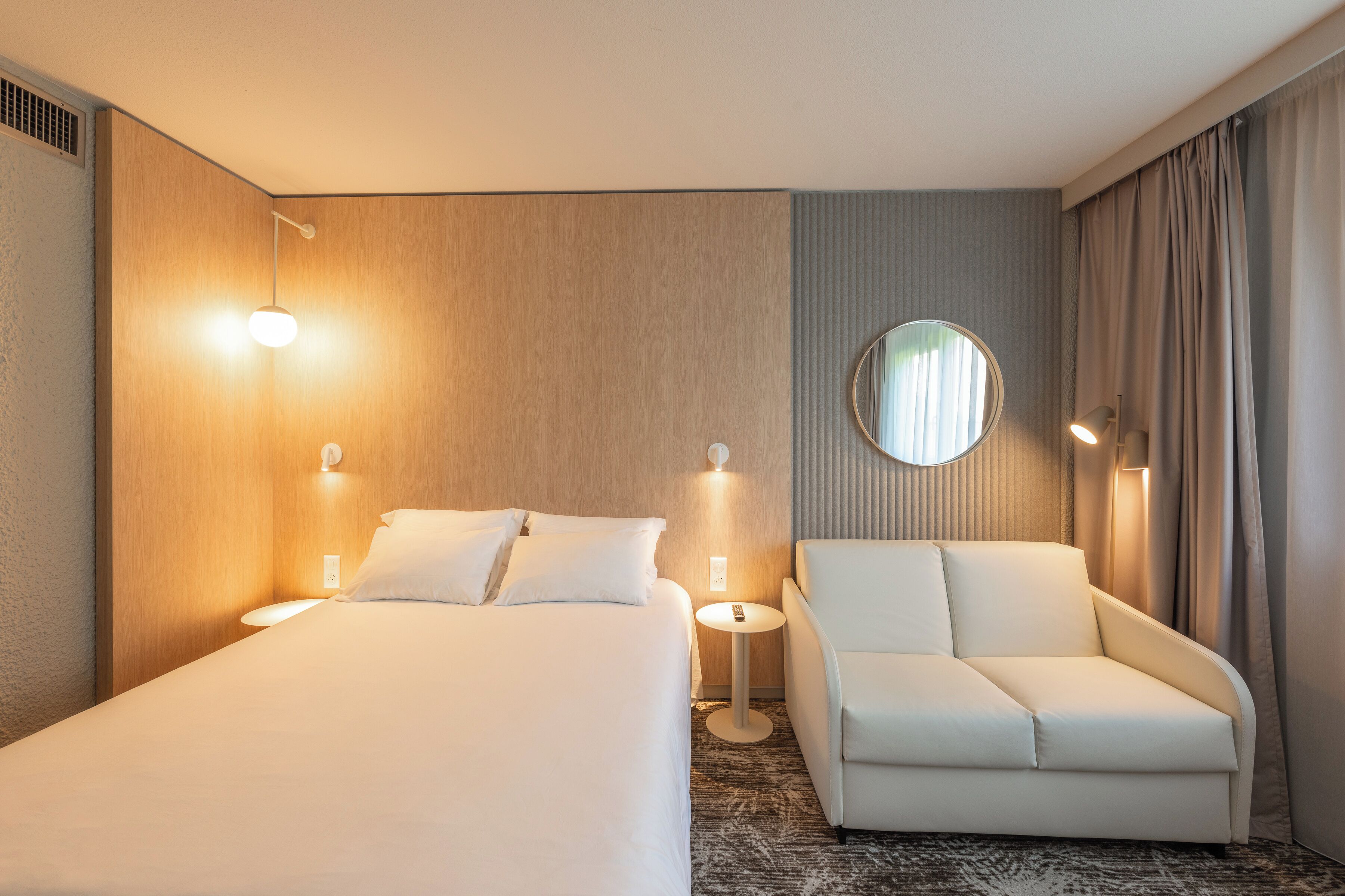 A hotel room with a bed, a sofa, a round mirror, and two wall-mounted lamps.