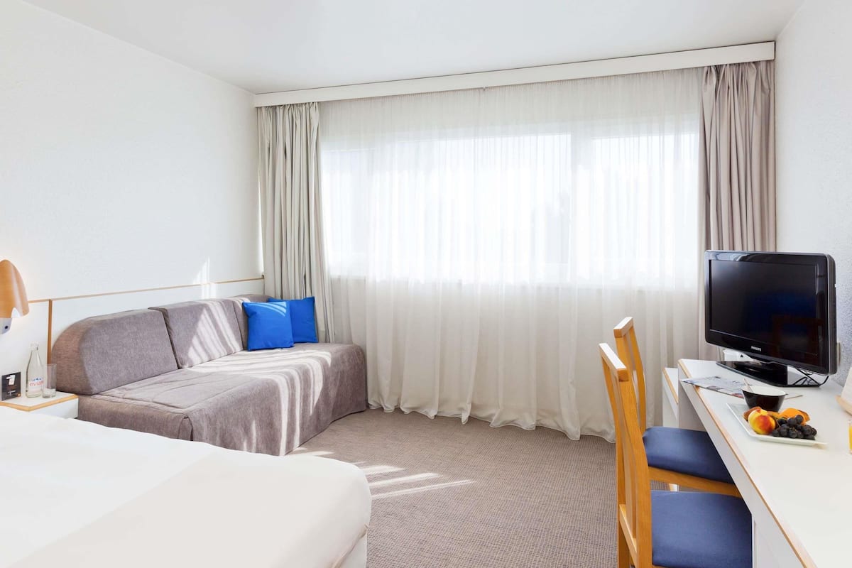 Ein Hotelzimmer mit zwei Betten, einem Schreibtisch mit Fernseher, einem Sessel und einem Fenster mit Vorhängen.