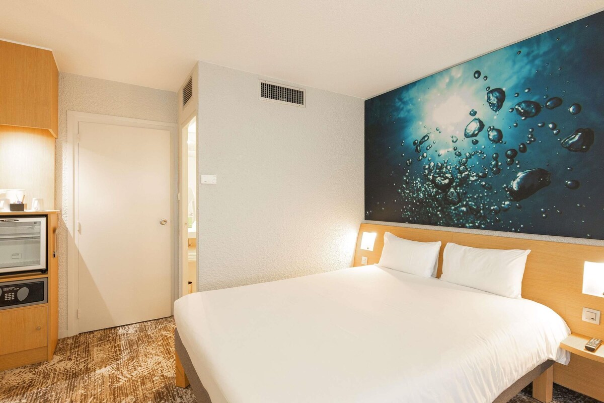 Habitación de hotel con una cama, televisor, un cuadro y piso alfombrado.