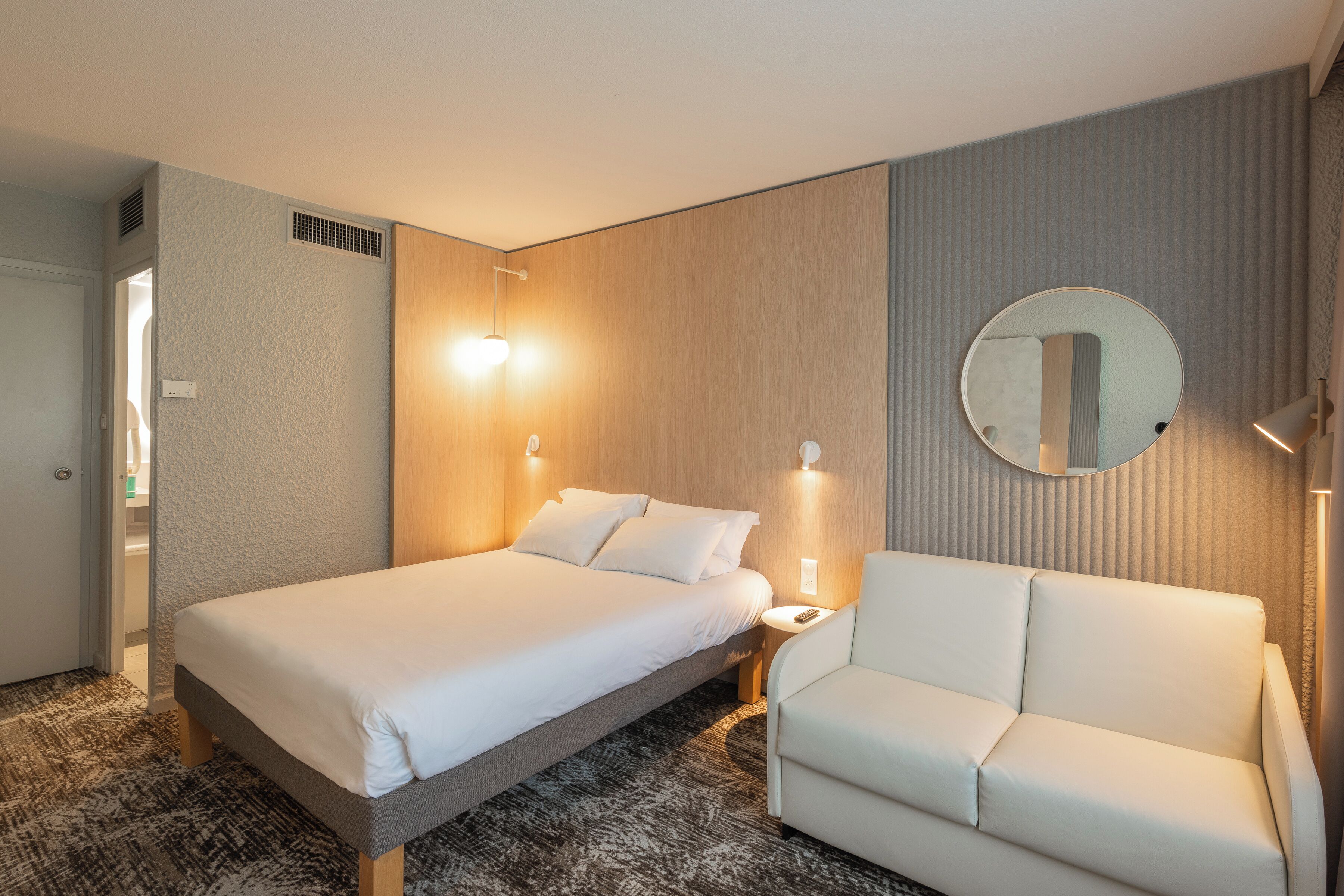 A hotel room with a bed, a chair, a bedside table, a mirror, and a wall-mounted light fixture.