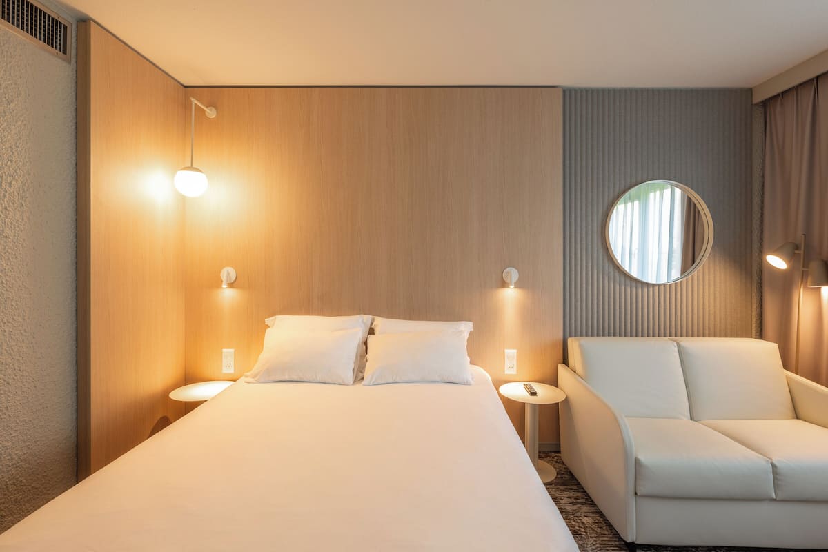 Ein Hotelzimmer mit einem Bett, zwei Nachttischen, einem runden Spiegel und einem weißen Sessel.