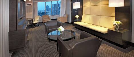 A modern hotel room with a large window, a sofa, two armchairs, a coffee table, and a TV.