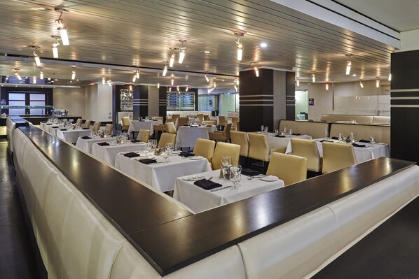 A dining area with multiple tables set for service, featuring white tablecloths, beige chairs, and overhead lighting.