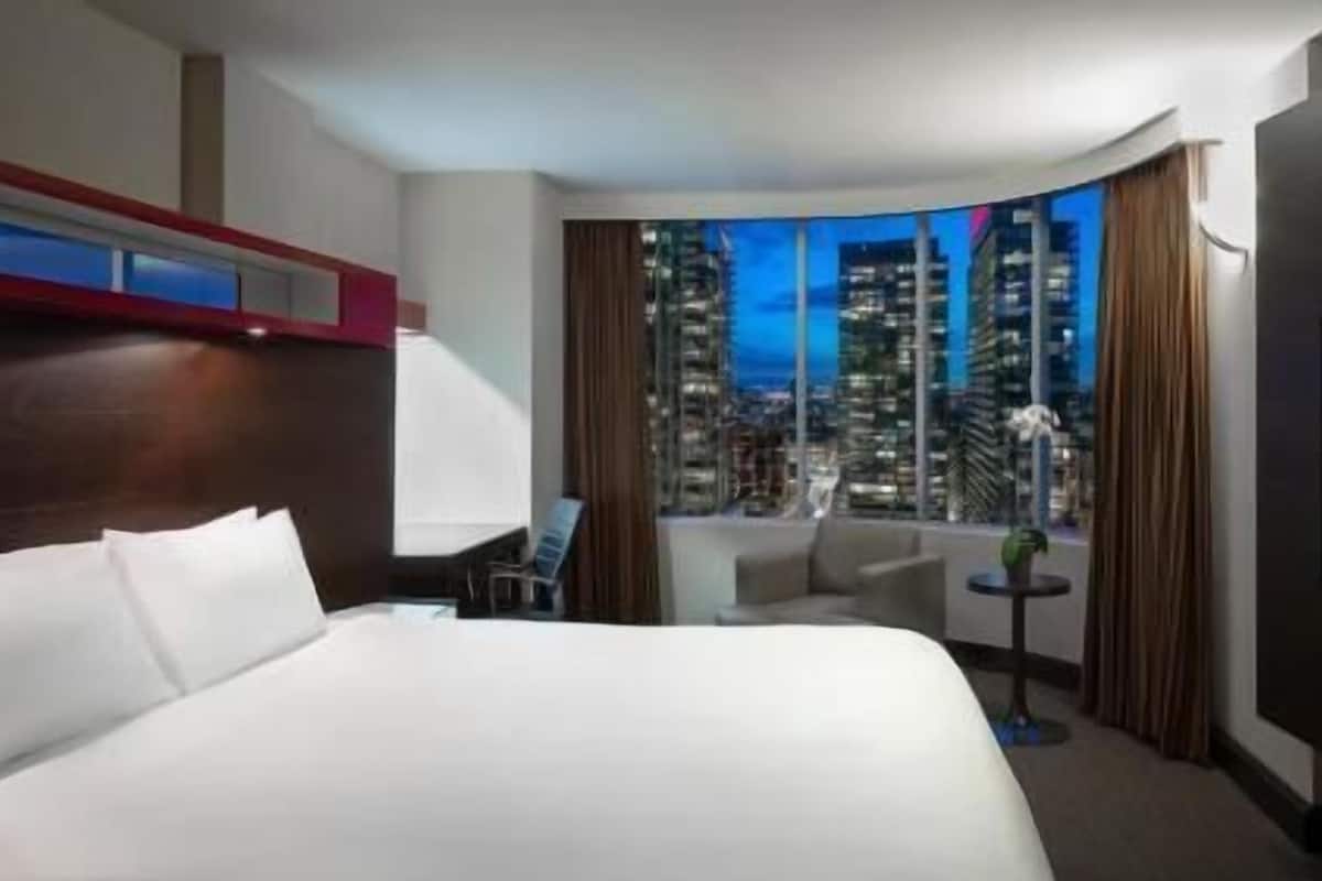 Habitación de hotel con una cama grande, televisión, zona de estar con vista a la ciudad iluminada por la noche y ventana con cortinas.