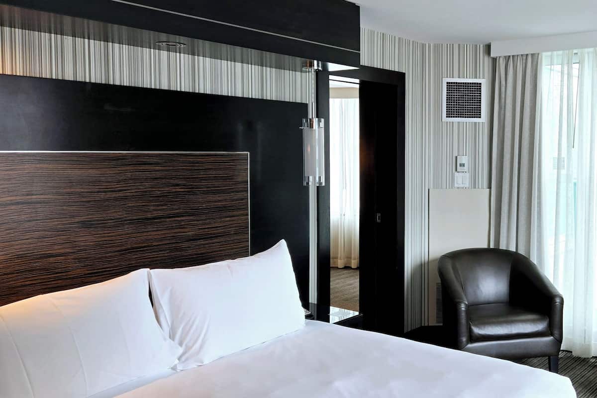 A modern hotel room with a large bed, a black leather chair, and a view of the city through the window.