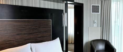 A modern hotel room with a large bed, a black leather chair, and a view of the city through the window.