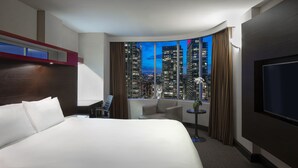A modern hotel room with a large bed, a seating area, a TV, and a view of a cityscape at dusk.