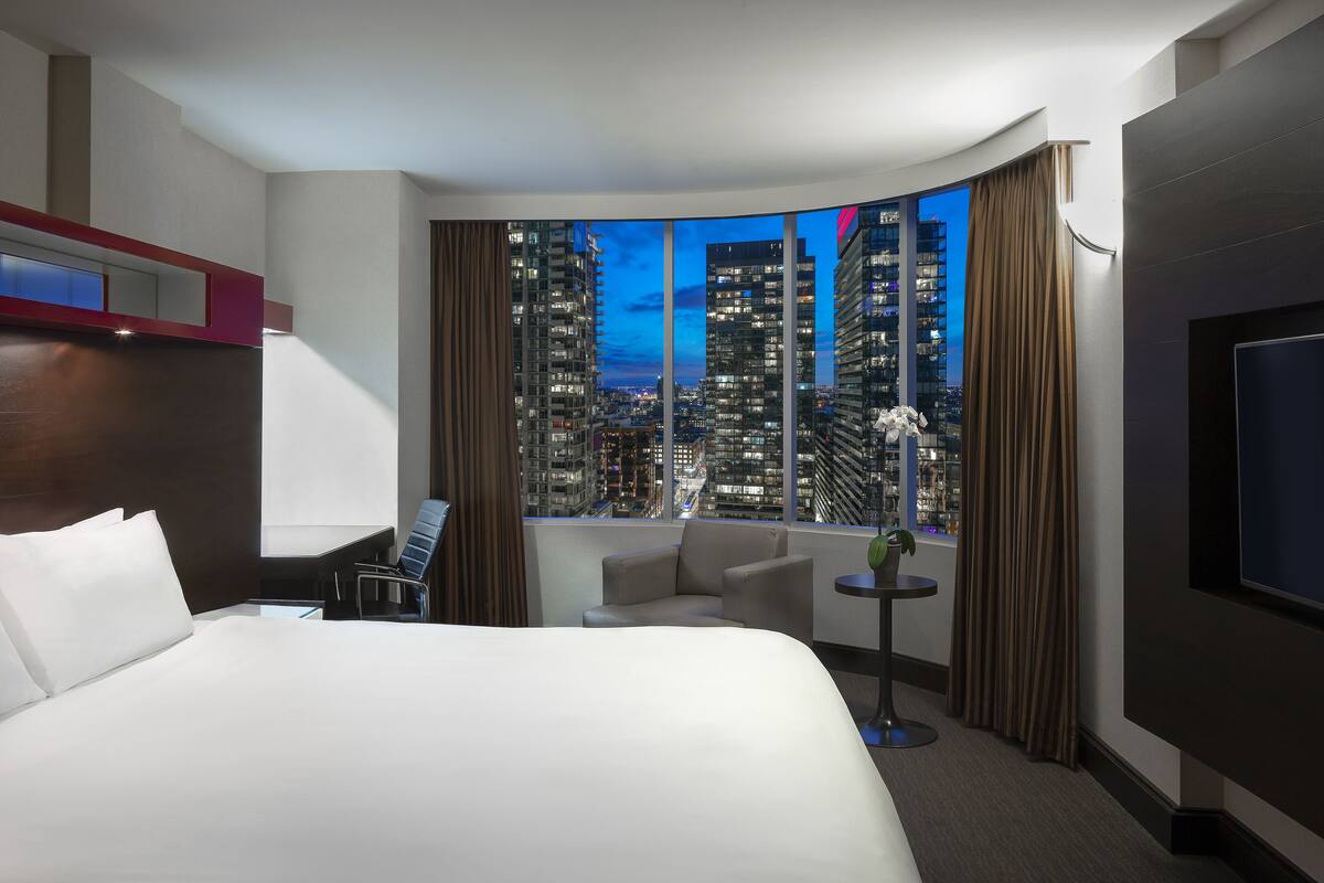 A modern hotel room with a large bed, a seating area, a TV, and a view of a cityscape at dusk.