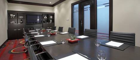 A conference room with a long table, chairs, a TV, and a window with blinds.