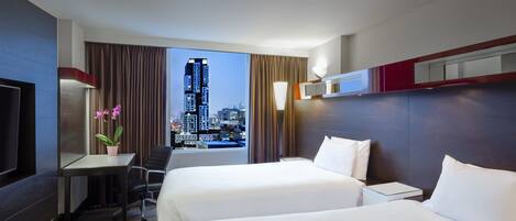 A hotel room with two beds, a desk with a chair, a TV, and a view of a cityscape.