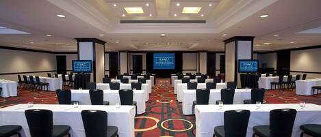 A conference room with rows of tables and chairs, a large screen at the front, and a carpeted floor.