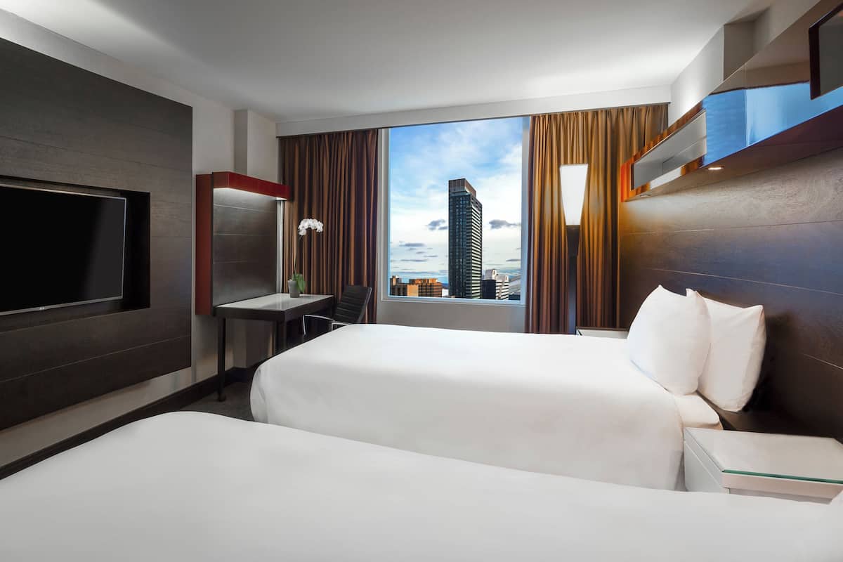 A hotel room with two beds, a desk, a TV, and a view of a cityscape.