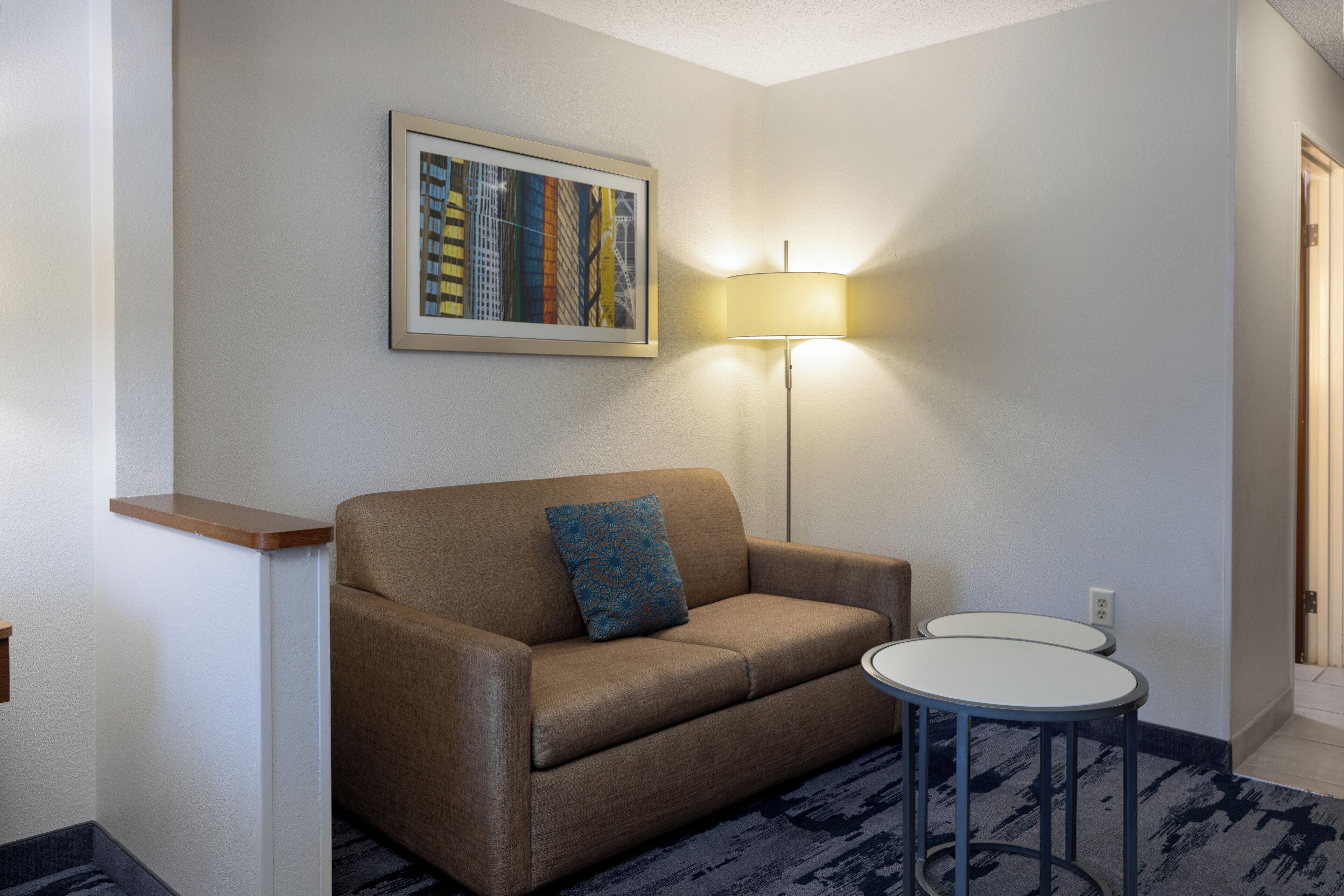 A hotel room with a brown sofa, a small round table, a lamp, and a framed picture on the wall.