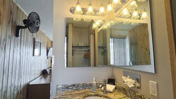 Un baño con tocador de madera, un espejo grande con iluminación y un ventilador, además de un cubo de basura.