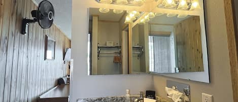 Un baño con tocador de madera, un espejo grande con iluminación y un ventilador, además de un cubo de basura.