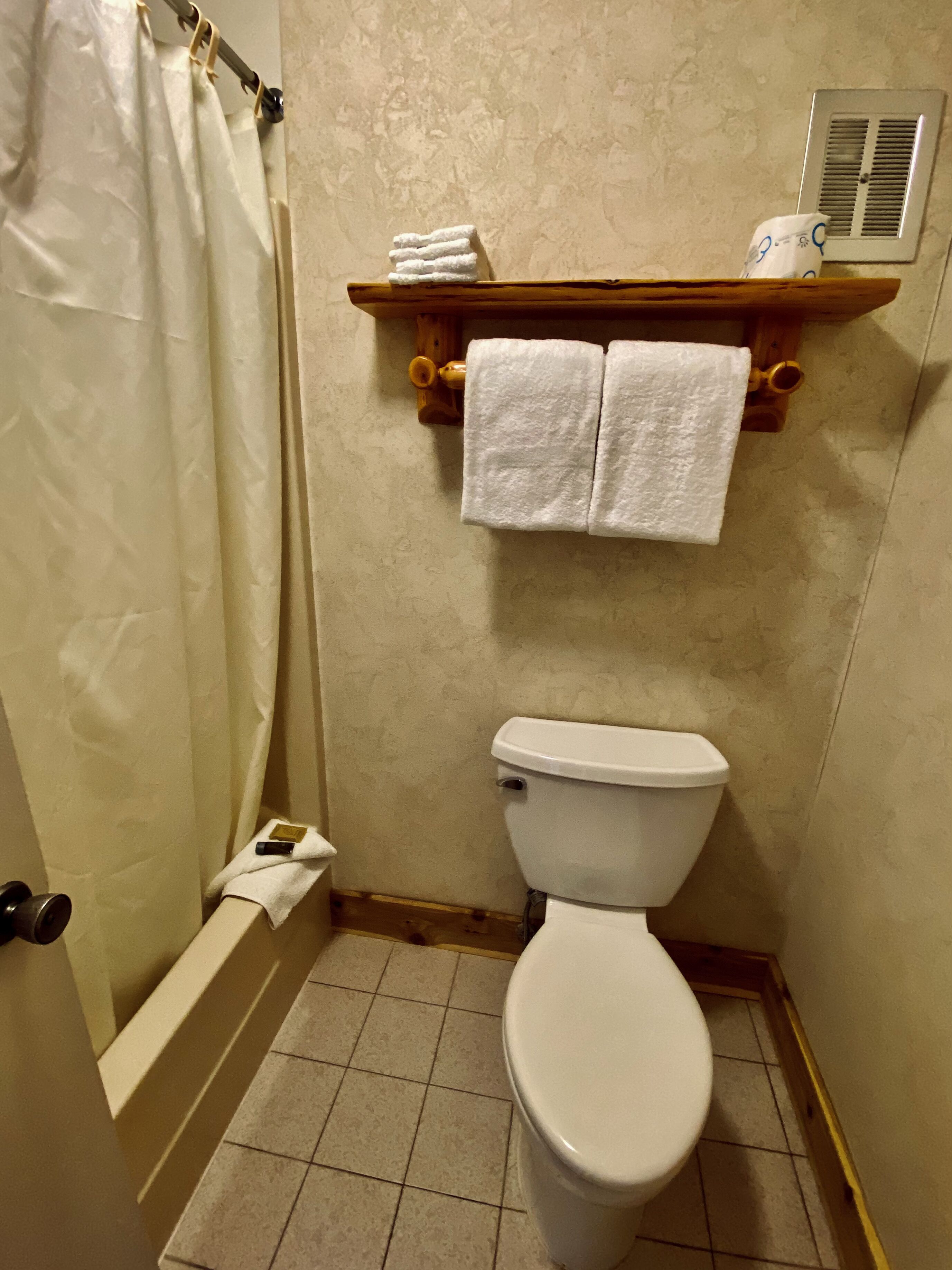 A bathroom with a toilet, a shower curtain, a towel rack with two towels, and a vent on the wall.